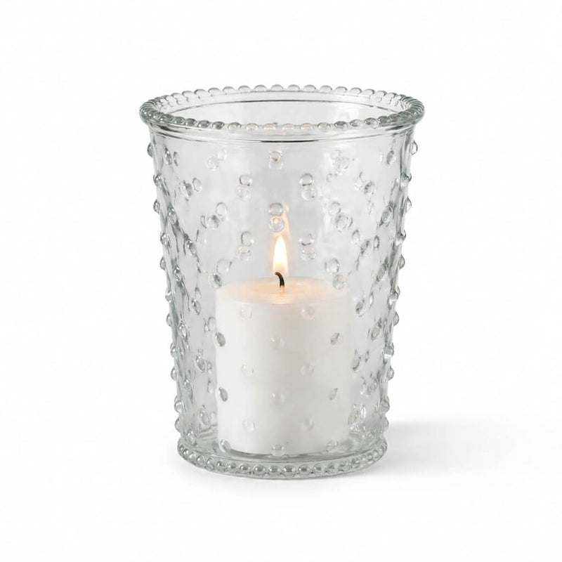 A lit white pillar candle inside a clear glass Hobnail Candle Holder on a white studio backdrop - thumbnail number 3