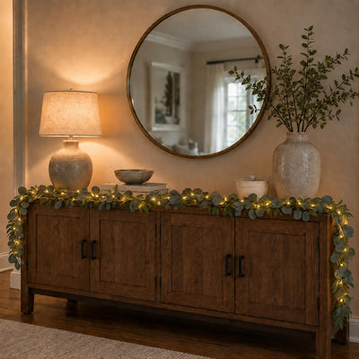 Eucalyptus garland with warm white LED lights draped over a wooden credenza under a round mirror - thumbnail number 8