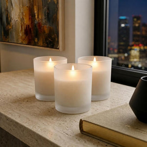 Three lit candles on a surface with a cityscape view through a window.