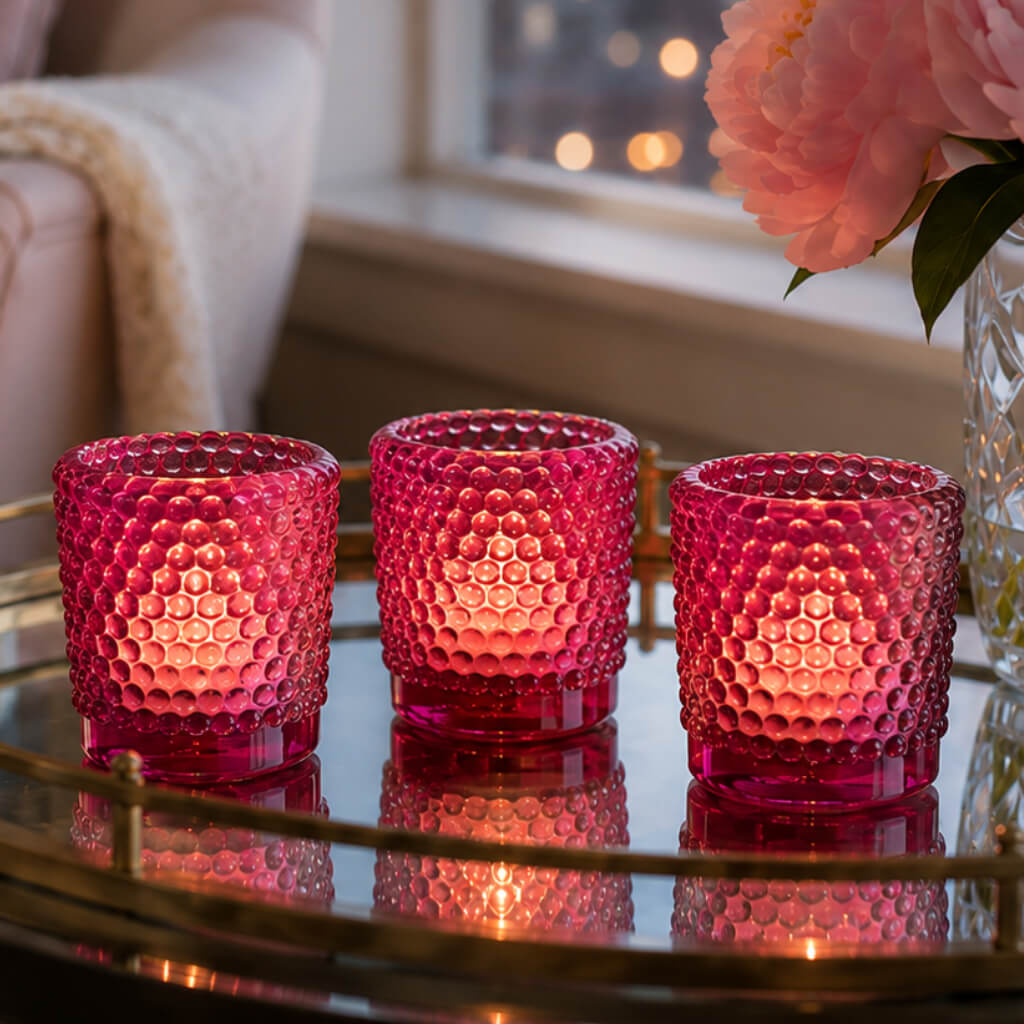 Three pink hobnail votive holders glowing on mirrored tray beside pink flowers