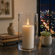 A lit white pillar candle inside a glass cylinder vase on a table by a window at night - thumbnail number 7