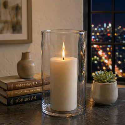 A lit white pillar candle inside a glass cylinder vase on a table by a window at night - thumbnail number 7
