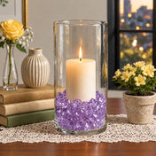 Lit white pillar candle in a glass cylinder filled with purple acrylic ice beads on a table - thumbnail number 9