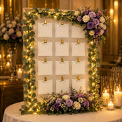 Eucalyptus garland lights frame a seating chart board decorated with purple and white flowers - thumbnail number 6