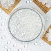 Top-down view of clear water gel beads filling a round glass bowl on a decorated table. - thumbnail number 1